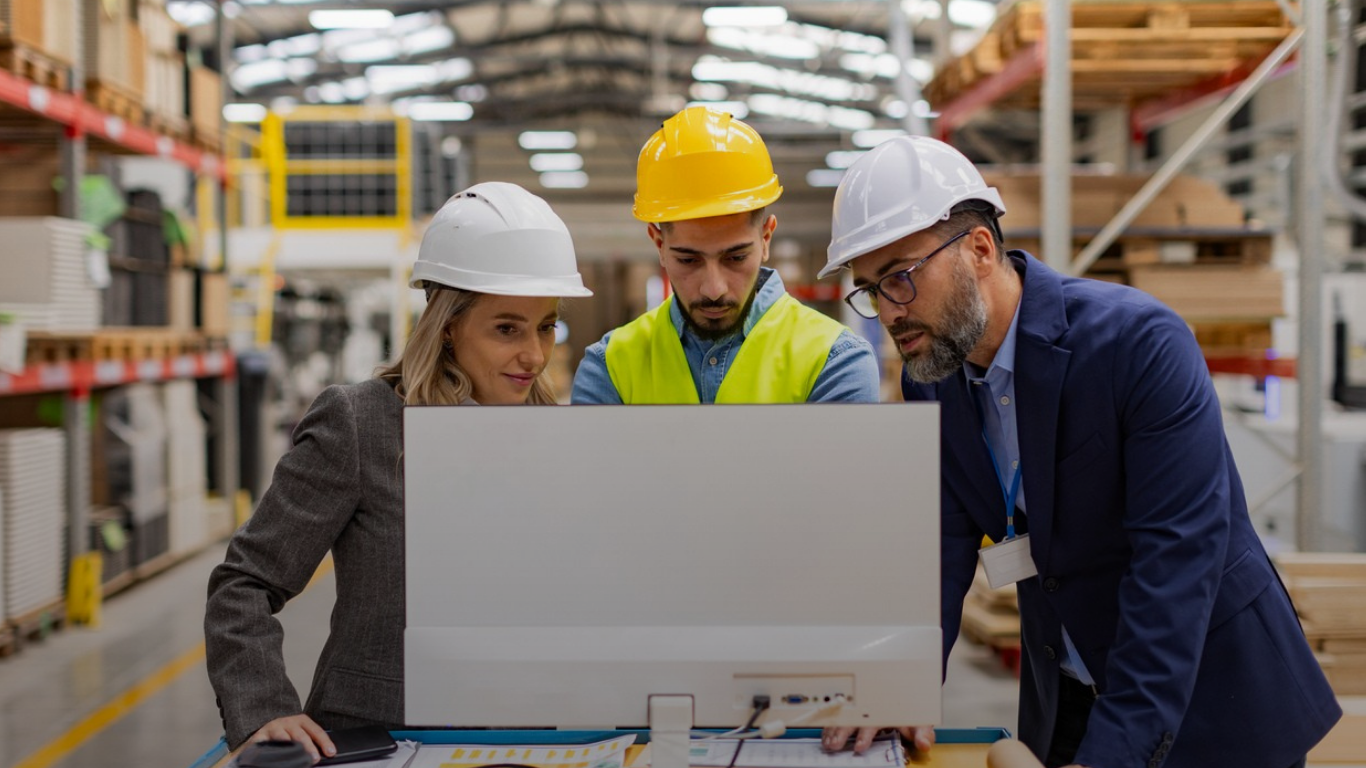 Three professionals in a factory reviewing data on a computer screen, discussing system performance and potential implementation